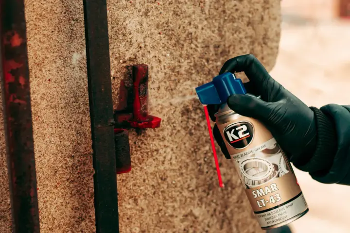 Ręka w czarnej rękawiczce aplikuje smar do przegubów wewnętrznych na zardzewiały element przy betonowym słupku.