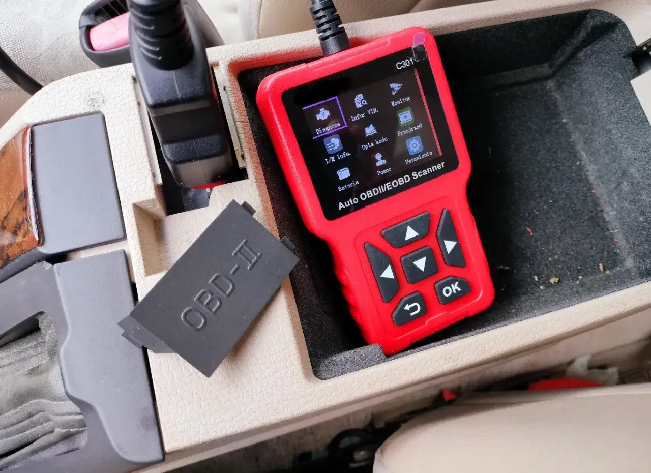 Czerwony skaner diagnostyczny Auto OBDII/EOBD Scanner leży w schowku samochodu obok zaślepki z napisem OBD-II.