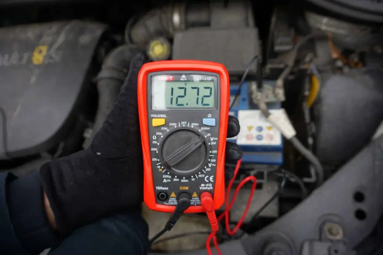 Ręka w czarnej rękawiczce trzyma multimetr pokazujący 12.72V, co świadczy o prawidłowym napięciu akumulatora.
